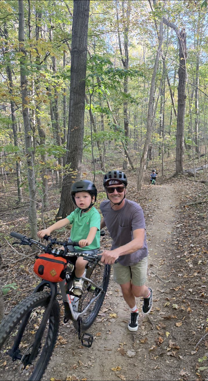 Kristian pushing his son Easton up a wooded trail on a bike